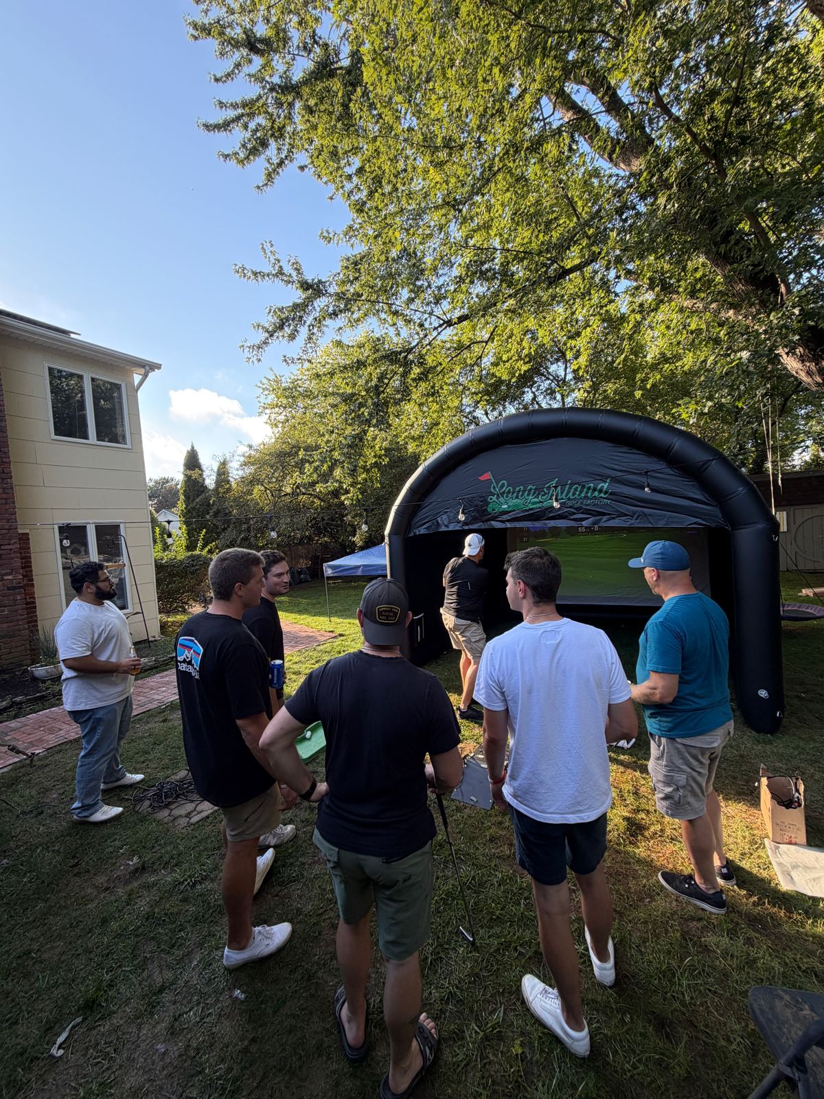 Guests gathered around the black Long Island Golf Factory inflatable golf simulator in a Long Island backyard on a sunny afternoon